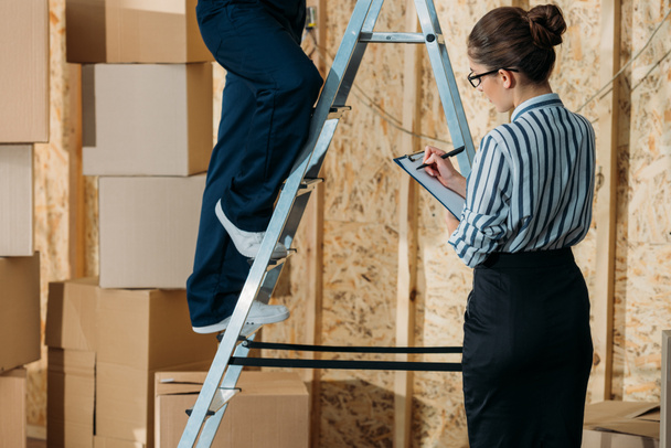 Businesswoman writing in clipboard while loader man standing on a ladder - Φωτογραφία, εικόνα