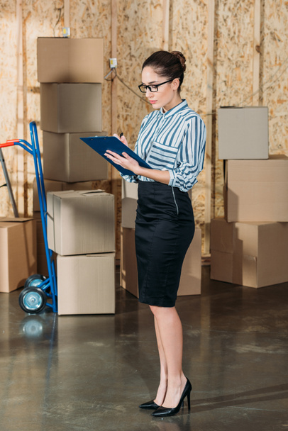 Young businesswoman writing in clipboard by stacks of delivery boxes - Photo, Image