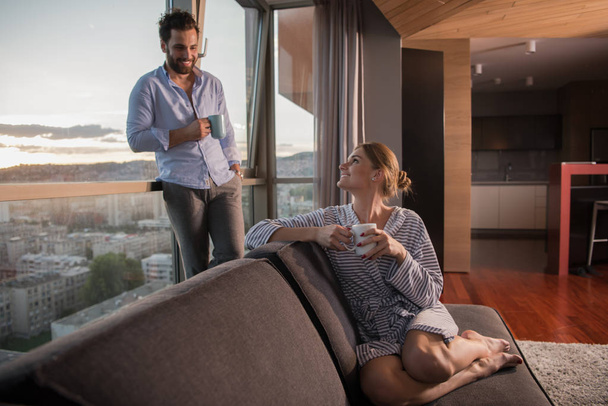 romantic happy young couple enjoying evening coffee and beautiful sunset landscape of the city while standing by the window - Фото, изображение