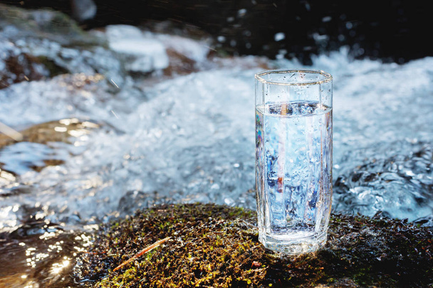 A transparent glass glass with drinking mountain water stands in the moss stone on sun beame against a background of a clean frost mountain river. The concept of drinking mountain drinking mineral - Fotografie, Obrázek