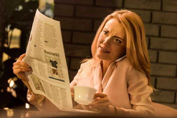 portrait of businesswoman with cup of coffee talking on smartphone while reading newspaper in cafe - Фото, изображение