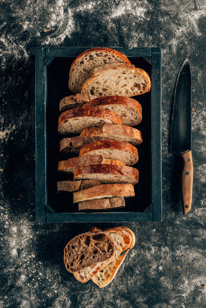 top view of arranged pieces of bread in wooden box and knife on dark tabletop with flour - Fotoğraf, Görsel