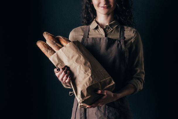 cropped shot of woman in apron holding french baguettes in paper bag in hands isolated on black - Fotografie, Obrázek