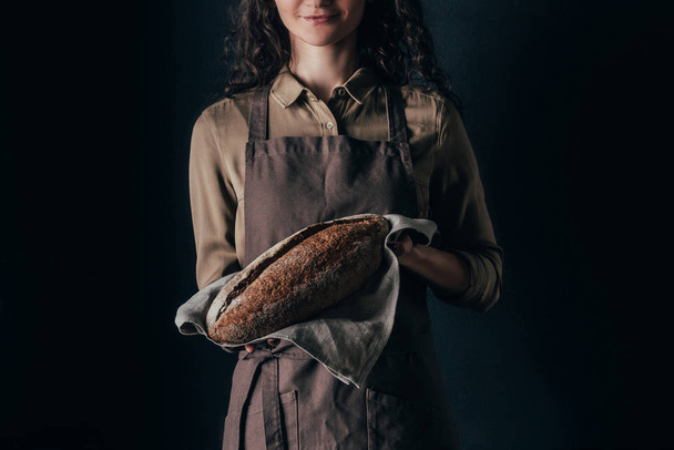partial view of woman in apron holding french baguette in hands isolated on black - Foto, Imagen