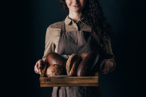 cropped shot of smiling woman holding wooden box with loafs of bread in hands isolated on black - Фото, изображение