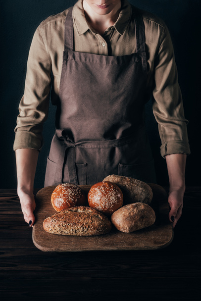partial view of woman holding loafs of bread on wooden cutting board isolated on black - Photo, image