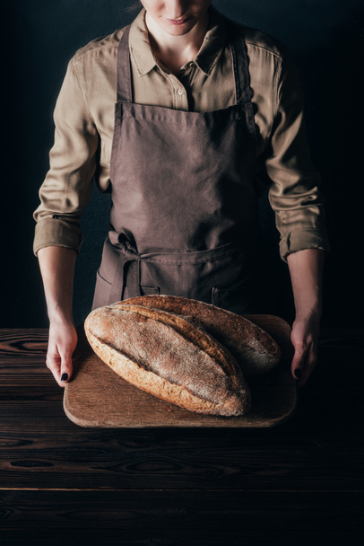 partial view of woman holding loafs of bread on wooden cutting board isolated on black - Foto, immagini