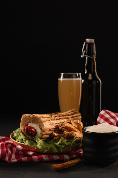νόστιμα Γαλλικά χοτ-ντογκ σε πιάτο με μπουκάλι και ποτήρι της μπύρας που απομονώνονται σε μαύρο - Φωτογραφία, εικόνα