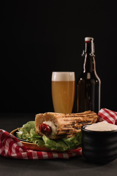 tasty french hot dog on plate with bottle and glass of beer isolated on black - Photo, Image