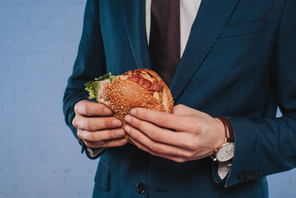 обрезанный снимок бизнесмена в костюме, держащего бургер
 - Фото, изображение