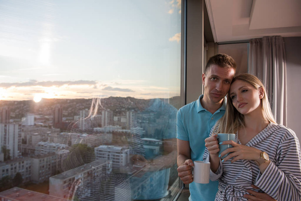 romantic happy young couple enjoying evening coffee and beautiful sunset landscape of the city while standing by the window - Foto, Bild