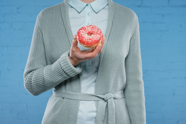 cropped shot of woman in sweater and shirt holding delicious glazed doughnut in front of blue brick wall - Фото, изображение