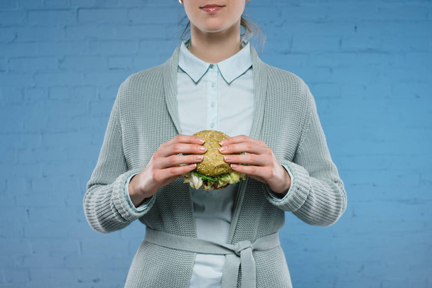cropped shot of young woman holding green vegan burger in front of blue brick wall - Photo, Image