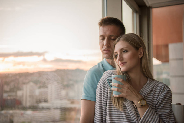 romantic happy young couple enjoying evening coffee and beautiful sunset landscape of the city while standing by the window - Photo, image