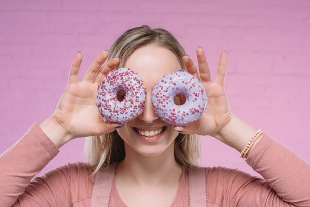 smiling young woman covering eyes with doughnuts in front of pink brick wall - Фото, изображение