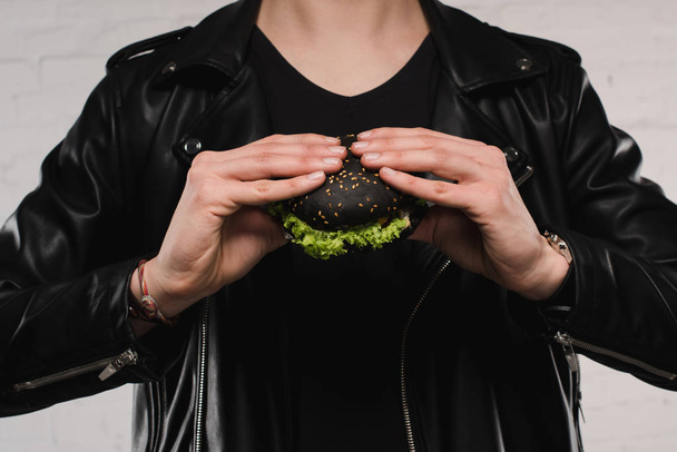 cropped shot of man in leather jacket holding black burger - Фото, изображение