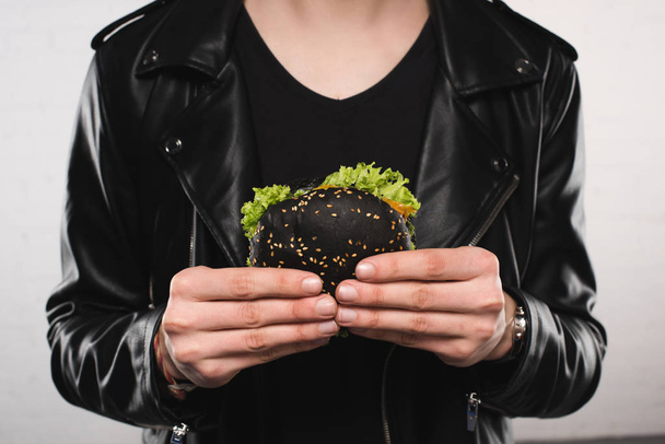 περικοπεί shot του ανθρώπου σε κομψό δερμάτινο μπουφάν κρατώντας μαύρα burger - Φωτογραφία, εικόνα
