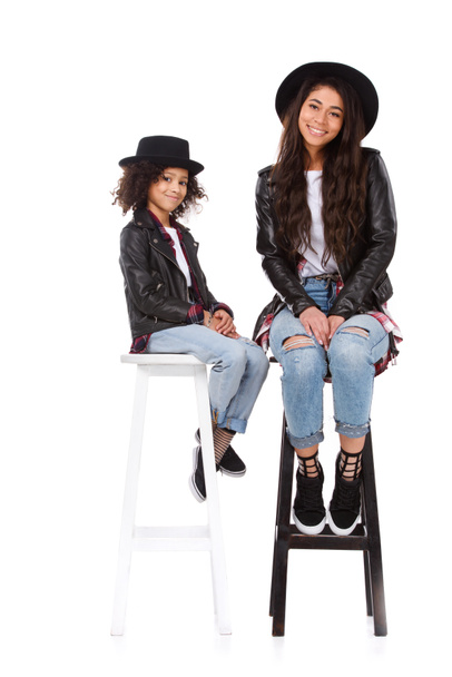 stilvolle Mutter und Tochter in Hüten und Lederjacken sitzen auf Stühlen isoliert auf weiß - Foto, Bild