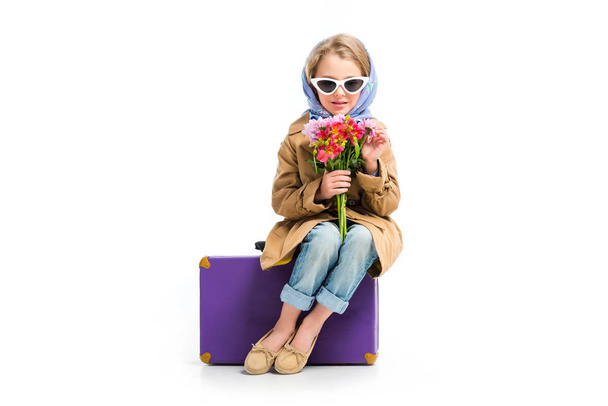 Stylish child in sunglasses and headscarf holding flowers while sitting on suitcase isolated on white - Photo, image