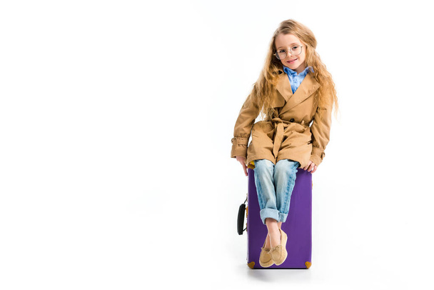 Stylish child in glasses and trench coat sitting on suitcase isolated on white - Fotografie, Obrázek