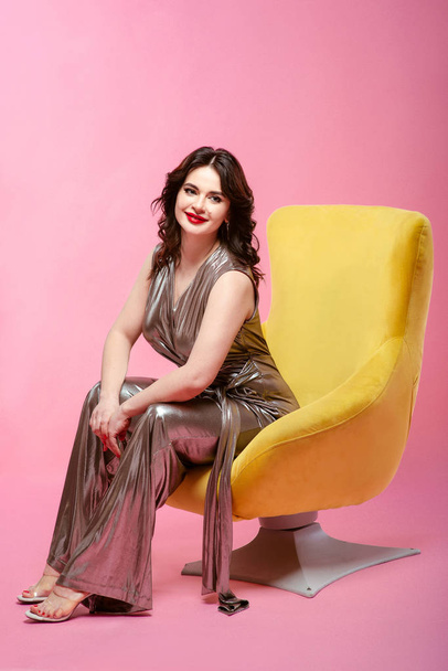 ピンクの背景に黄色のスタイリッシュなアームチェアに座って銀ディスコ オーバー オールでブルネットのはかなり魅力的な若い女性の肖像画 - 写真・画像