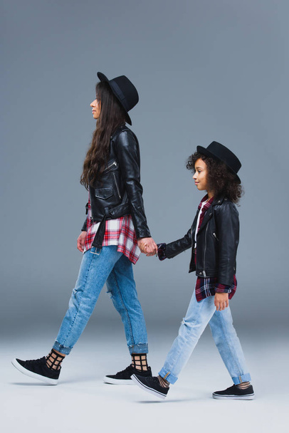 side view of stylish mother and daughter walking together and holding hands on grey - Fotoğraf, Görsel