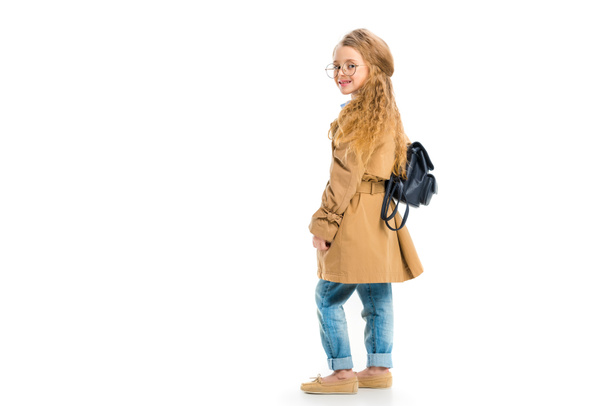 Rear view of smiling child in glasses wearing trench coat and holding bag isolated on white - Foto, Imagem