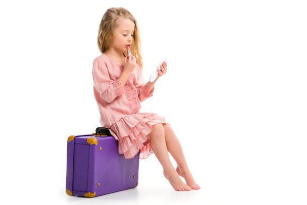 Side view of stylish child sitting on suitcase and doing makeup by lipstick isolated on white - Fotó, kép