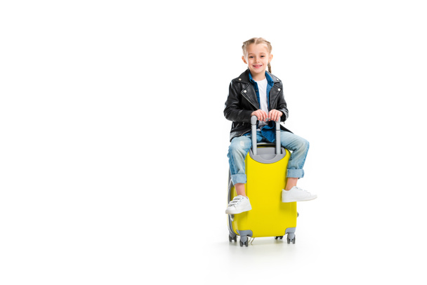 Piccolo turista con le trecce che indossa giacca di pelle e seduto su valigia a rotelle isolato su bianco
 - Foto, immagini