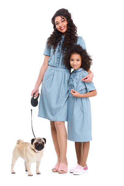 jonge moeder en dochter in soortgelijke jurken met pug aan leiband geïsoleerd op wit - Foto, afbeelding