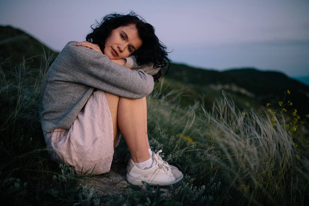 συλλογισμένος όμορφη μελαχρινή γυναίκα κάθεται στο γρασίδι του λόφου πάνω από τη θάλασσα. το κορίτσι χαλάρωση στους εξωτερικούς χώρους.   - Φωτογραφία, εικόνα