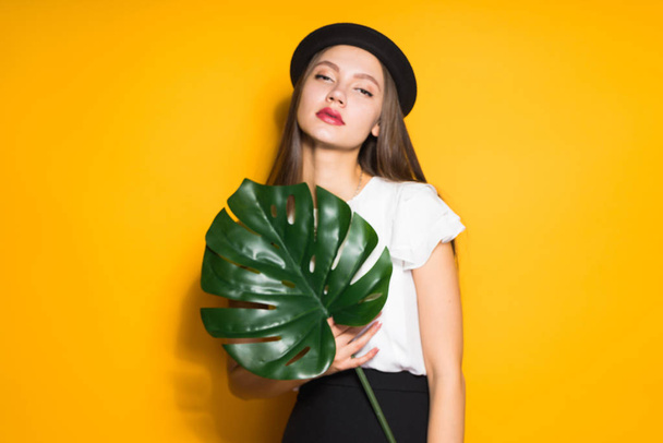 attractive young woman in fashion hat posing on yellow background, holding green leaf - Foto, imagen