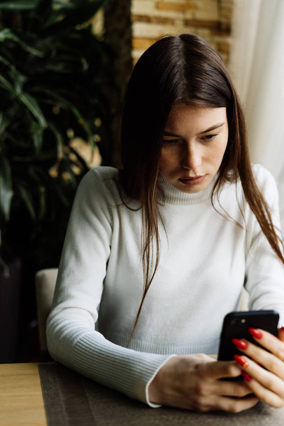 roztomilá mladá dívka s tmavými vlasy a v bílých svetr sedí v kavárně a dívá do smartphone - Fotografie, Obrázek