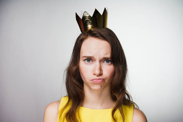 disgruntled young woman in a yellow dress looks at the camera, on her head a golden crown - Photo, Image