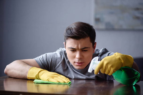 handsome man dusting table in living room with spray bottle and rag - Photo, Image