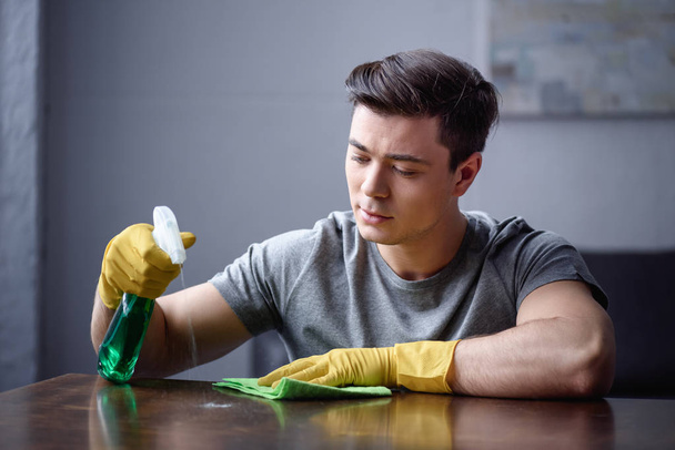 man cleaning table in living room with spray bottle and rag - Foto, Bild