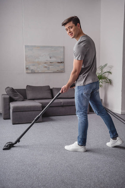 handsome man cleaning carpet with vacuum cleaner and looking at camera in living room - Photo, Image