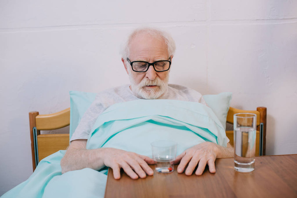 Старший пацієнт у клінічному ліжку дивиться на свої ліки
 - Фото, зображення