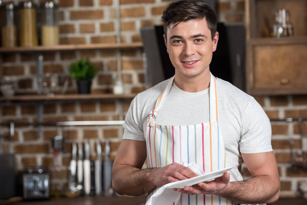 hombre guapo limpiando plato con toalla y mirando a la cámara en la cocina
 - Foto, Imagen