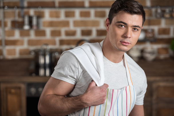 handsome man in apron with towel on shoulder looking at camera at kitchen - Foto, Bild