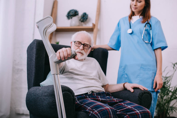 Senior man with crutch sitting in chair by female nurse - Zdjęcie, obraz