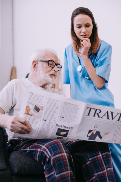 Pielęgniarka stojący przez starszego mężczyzny czytanie gazety - Zdjęcie, obraz