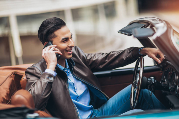 Fashion man sitting in luxury retro cabriolet car outdoors using phone - Foto, afbeelding