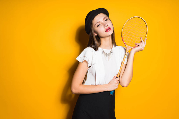 attractive girl in a black hat posing on a yellow background, holding a tennis racket - Fotó, kép