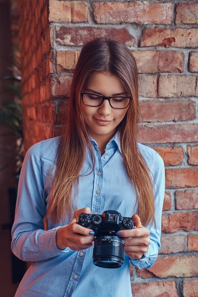 Portret młodego fotografa kobiece w okulary i niebieska koszula, opierając się na cegły ściany w pokoju z loft design. - Zdjęcie, obraz