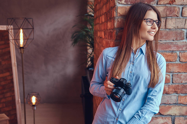 Portrait d'une jeune photographe en lunettes et chemise bleue souriante appuyée contre un mur de briques dans une pièce au design loft
. - Photo, image