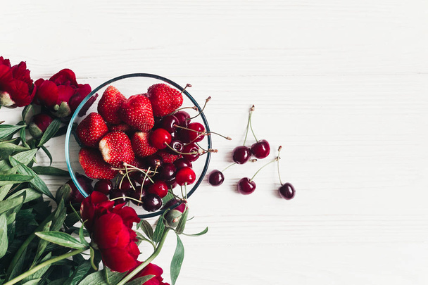 Sommerflache Liege mit frischen Kirschen und Erdbeeren in stilvoller Glasschale und Pfingstrosen mit Blättern auf weißem Holzgrund. - Foto, Bild