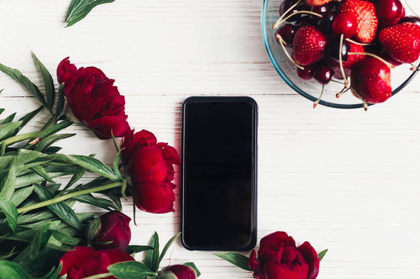 telefoon met leeg scherm en mooie rode pioenrozen, aardbeien en kersen op witte houten achtergrond plat leggen.  - Foto, afbeelding