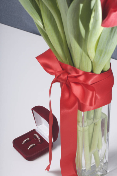 Gifts for loved ones. A bouquet of red tulips stands in a glass vase, tied with scarlet ribbon. Nearby is a red velvet box with a ring and earrings. - Photo, image