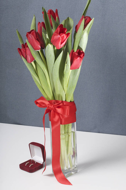 Gifts for loved ones. A bouquet of red tulips stands in a glass vase, tied with scarlet ribbon. Nearby is a red velvet box with a ring and earrings. - Valokuva, kuva